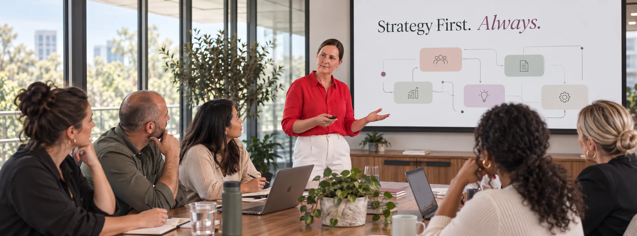 Kylie presenting to a team around a boardroom table, with a Strategy First slide on the screen behind her.