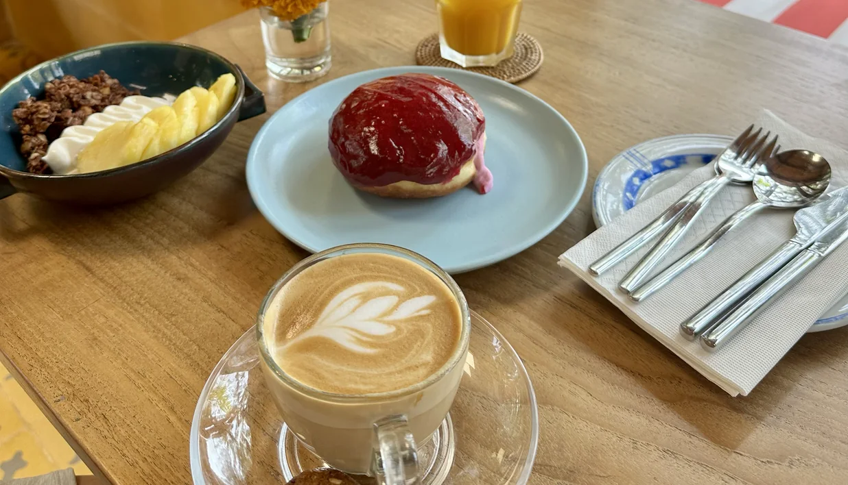 A brunch table with a latte, a jam doughnut, a fruit and granola bowl and juice, beside an open window.